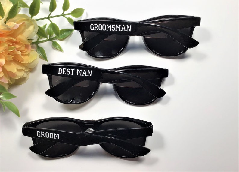 May include: Three pairs of black sunglasses with white text on the frames. The sunglasses say "Groom", "Best Man", and "Groomsman".
