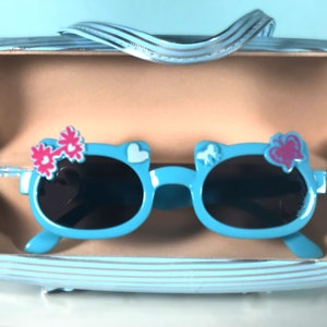 May include: Light blue sunglasses with black lenses, shaped like a cat's face, decorated with pink flower and heart accents. The sunglasses are in a matching blue striped case with a handle.