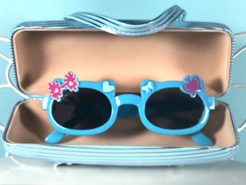 May include: Light blue sunglasses with black lenses, shaped like a cat's face, decorated with pink flower and heart accents. The sunglasses are in a matching blue striped case with a handle.