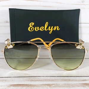 Op de afbeelding: Gouden aviator zonnebril met een groene lens met verloop en gele vlinderaccenten op de pootjes. De zonnebril ligt op een houten oppervlak met een zwarte etui waarop "Evelyn" in gele letters staat.