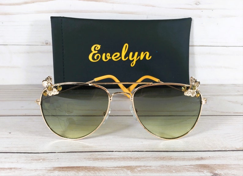 Op de afbeelding: Gouden aviator zonnebril met een groene lens met verloop en gele vlinderaccenten op de pootjes. De zonnebril ligt op een houten oppervlak met een zwarte etui waarop "Evelyn" in gele letters staat.