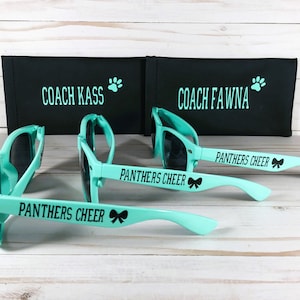 Puede incluir: Tres pares de gafas de sol color verde azulado claro con lentes negras y texto negro PANTHERS CHEER con un icono de lazo en las patillas. Detr&aacute;s, dos estuches rectangulares negros con texto en verde azulado COACH KASS y COACH FAWNA.