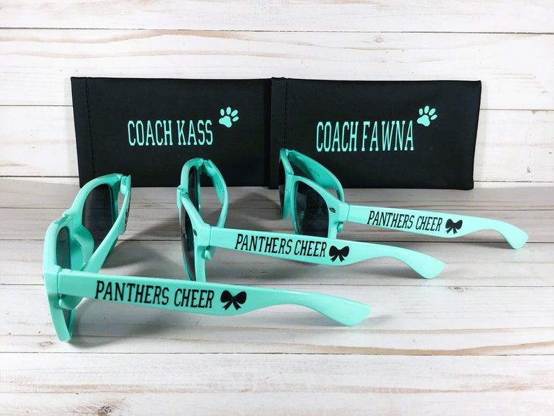 Puede incluir: Tres pares de gafas de sol color verde azulado claro con lentes negras y texto negro PANTHERS CHEER con un icono de lazo en las patillas. Detr&aacute;s, dos estuches rectangulares negros con texto en verde azulado COACH KASS y COACH FAWNA.