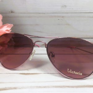May include: Aviator sunglasses with a metallic rose gold frame and tinted lenses. The right lens features the word Victoria printed in white script. The thin metal temples have pink plastic tips, and the nose pads are clear and adjustable.