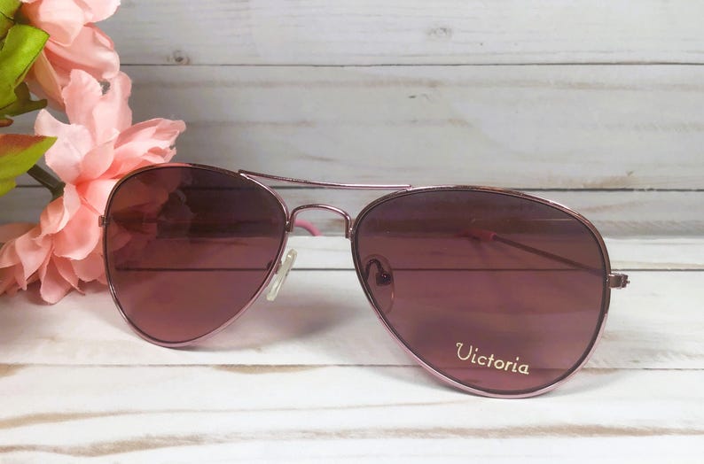 May include: Aviator sunglasses with a metallic rose gold frame and tinted lenses. The right lens features the word Victoria printed in white script. The thin metal temples have pink plastic tips, and the nose pads are clear and adjustable.