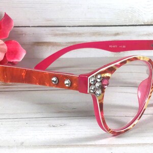 May include: Reading glasses with a bright pink and orange marbled frame, featuring clear lenses and decorative clear rhinestones embedded along the temples and front corners. The inner temple arm displays the text RD-977 +1.50.