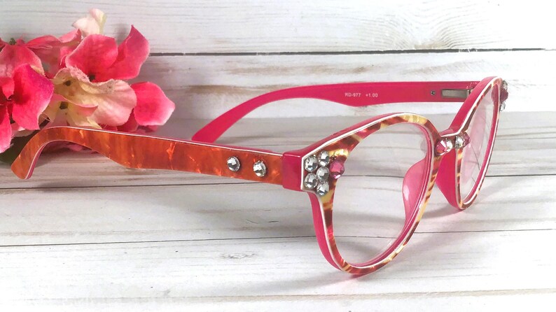 May include: Reading glasses with a bright pink and orange marbled frame, featuring clear lenses and decorative clear rhinestones embedded along the temples and front corners. The inner temple arm displays the text RD-977 +1.50.