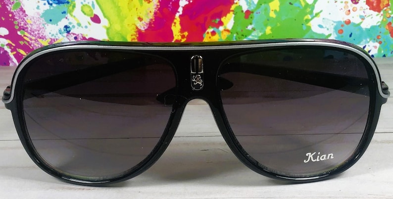 Puede incluir: Gafas de sol de aviador negras con ribete plateado y lentes degradadas. Las gafas de sol tienen un emblema decorativo en el puente y el nombre "Kian" impreso en la lente.