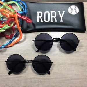 以下が含まれることがあります： 黒い丸いサングラスが2組と、白い文字で「RORY」と白の野球のグラフィックが書かれた黒いケース。