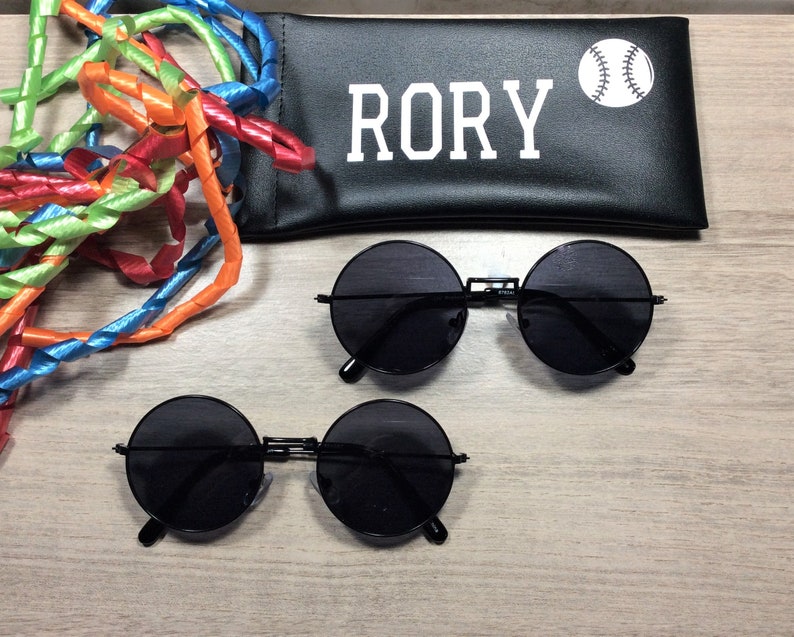 以下が含まれることがあります： 黒い丸いサングラスが2組と、白い文字で「RORY」と白の野球のグラフィックが書かれた黒いケース。