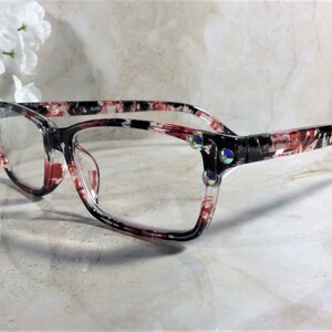 Pu&ograve; includere: Occhiali rettangolari con montatura a fantasia nera, rossa e bianca. Gli occhiali hanno lenti trasparenti e sono decorati con strass iridescenti sugli angoli. Lo stile generale &egrave; moderno.