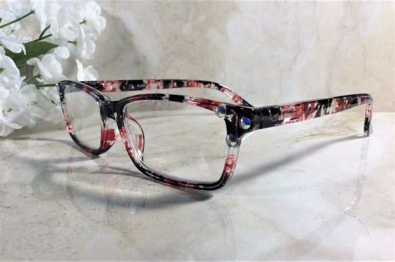 Pu&ograve; includere: Occhiali rettangolari con montatura a fantasia nera, rossa e bianca. Gli occhiali hanno lenti trasparenti e sono decorati con strass iridescenti sugli angoli. Lo stile generale &egrave; moderno.