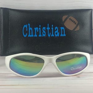 Op de afbeelding: Witte zonnebril met iriserende glazen met de naam Christian op het rechterglas gedrukt. De zonnebril staat voor een zwart rechthoekig etui met de naam Christian en een bruine afbeelding van een American football erop gedrukt.