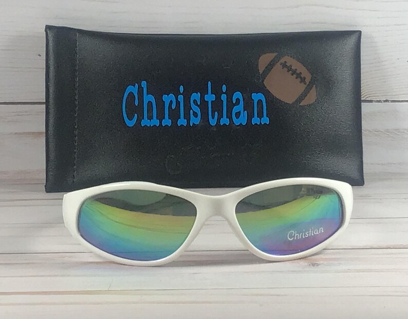 Op de afbeelding: Witte zonnebril met iriserende glazen met de naam Christian op het rechterglas gedrukt. De zonnebril staat voor een zwart rechthoekig etui met de naam Christian en een bruine afbeelding van een American football erop gedrukt.