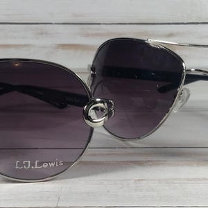May include: Two pairs of aviator sunglasses with dark purple lenses and silver frames. The sunglasses have a double bridge and the text "L.J. Lewis" and "S. Lewis" are printed on the lenses. The sunglasses are displayed on a light wood surface.