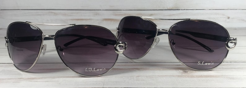 May include: Two pairs of aviator sunglasses with dark purple lenses and silver frames. The sunglasses have a double bridge and the text "L.J. Lewis" and "S. Lewis" are printed on the lenses. The sunglasses are displayed on a light wood surface.
