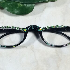 May include: Black reading glasses with a decorative floral pattern in yellow and teal. The top of the frame is embellished with a row of green rhinestones. The glasses have a classic half-frame design.