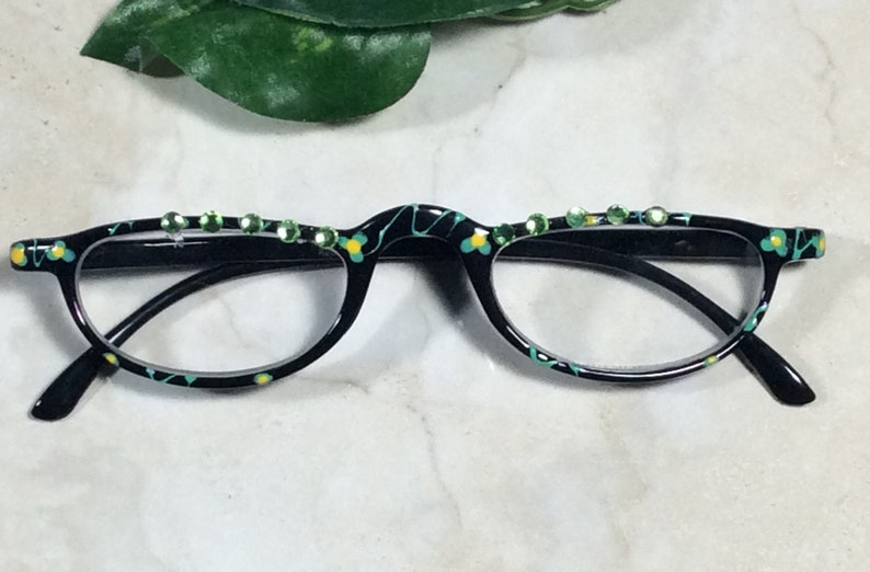 May include: Black reading glasses with a decorative floral pattern in yellow and teal. The top of the frame is embellished with a row of green rhinestones. The glasses have a classic half-frame design.