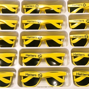 Puede incluir: Una colecci&oacute;n de gafas de sol de color amarillo brillante dispuestas en una bandeja de pl&aacute;stico blanca. Cada par tiene lentes negras y la palabra Hatters impresa en la patilla, acompa&ntilde;ada de un peque&ntilde;o gr&aacute;fico de un bal&oacute;n de f&uacute;tbol.