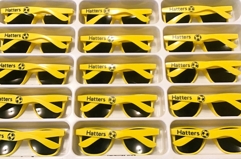 Puede incluir: Una colecci&oacute;n de gafas de sol de color amarillo brillante dispuestas en una bandeja de pl&aacute;stico blanca. Cada par tiene lentes negras y la palabra Hatters impresa en la patilla, acompa&ntilde;ada de un peque&ntilde;o gr&aacute;fico de un bal&oacute;n de f&uacute;tbol.