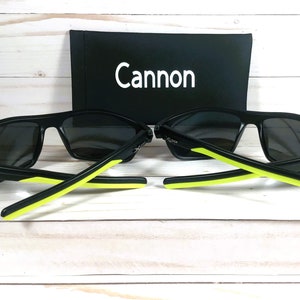 Peut inclure: Deux paires de lunettes de soleil noires avec des verres gris et des accents vert n&eacute;on sur les branches. Les lunettes sont pr&eacute;sent&eacute;es avec un &eacute;tui noir portant le mot "Cannon" en blanc.