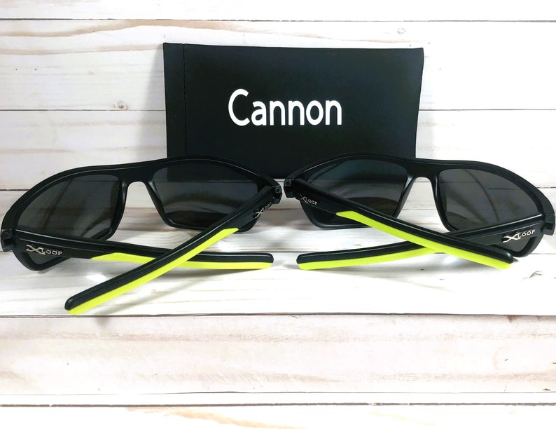 Peut inclure: Deux paires de lunettes de soleil noires avec des verres gris et des accents vert n&eacute;on sur les branches. Les lunettes sont pr&eacute;sent&eacute;es avec un &eacute;tui noir portant le mot "Cannon" en blanc.