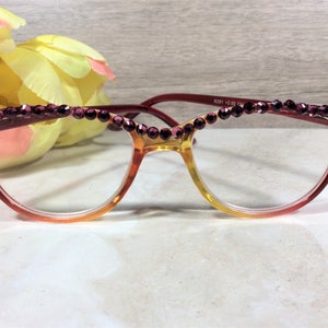 May include: Reading glasses with a gradient frame, transitioning from red to yellow. The top of the frame is adorned with a row of purple rhinestones. The glasses have clear lenses and are set against a soft background.