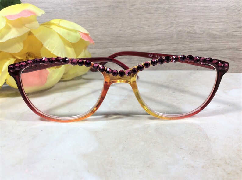 May include: Reading glasses with a gradient frame, transitioning from red to yellow. The top of the frame is adorned with a row of purple rhinestones. The glasses have clear lenses and are set against a soft background.