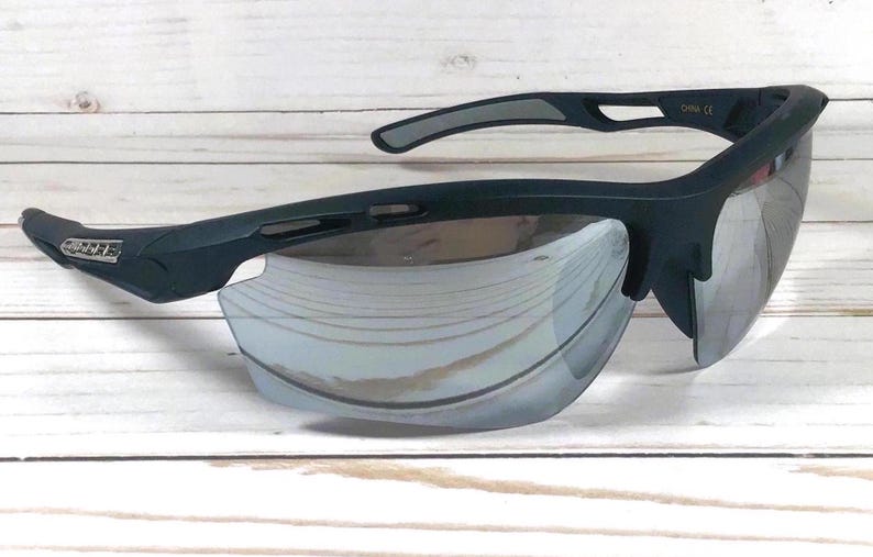 May include: Matte black sports sunglasses featuring a wraparound frame design with silver mirrored lenses. The temples display a small rectangular logo plate on the side, and the frame includes ventilation cutouts along the top brow bar.