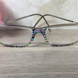 May include: Silver-framed eyeglasses with clear lenses. The frames are adorned with colorful rhinestones in shades of pink, green, and blue. The glasses are set against a light wooden surface, with a pink flower in the background.