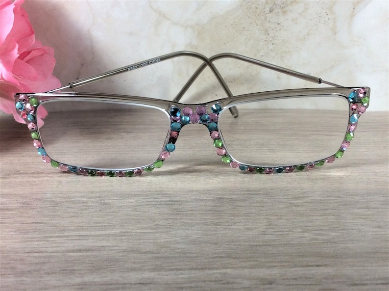 May include: Silver-framed eyeglasses with clear lenses. The frames are adorned with colorful rhinestones in shades of pink, green, and blue. The glasses are set against a light wooden surface, with a pink flower in the background.