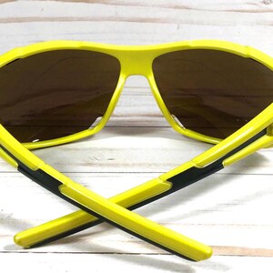 May include: Yellow sunglasses with black accents and brown lenses. The sunglasses have a sporty design and are ideal for outdoor activities.