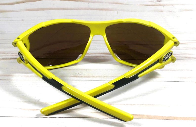 May include: Yellow sunglasses with black accents and brown lenses. The sunglasses have a sporty design and are ideal for outdoor activities.