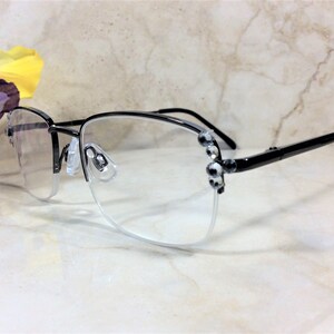 May include: Black and silver-toned eyeglasses with a half-rim design. The frames feature decorative embellishments on the temples. The lenses are clear, and the nose pads are transparent. A yellow and purple flower is in the background.