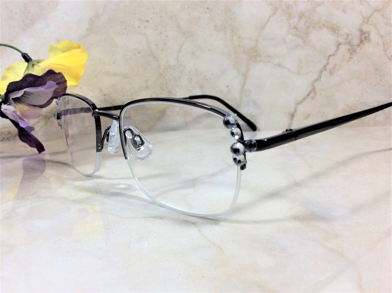 May include: Black and silver-toned eyeglasses with a half-rim design. The frames feature decorative embellishments on the temples. The lenses are clear, and the nose pads are transparent. A yellow and purple flower is in the background.