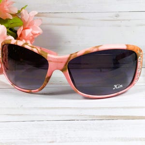 May include: Pink and brown camouflage sunglasses with rhinestones on the arms. The sunglasses have a dark lens and the word "Kim" is printed on the right lens.