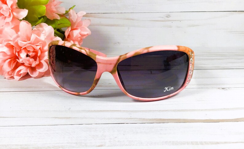 May include: Pink and brown camouflage sunglasses with rhinestones on the arms. The sunglasses have a dark lens and the word "Kim" is printed on the right lens.