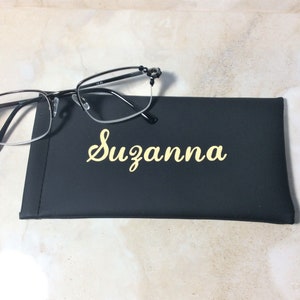 May include: Black-framed spectacles sit on a black case, personalised with the name "Suzanna" in gold script. The glasses have a half-rim design. The case is a dark rectangle.