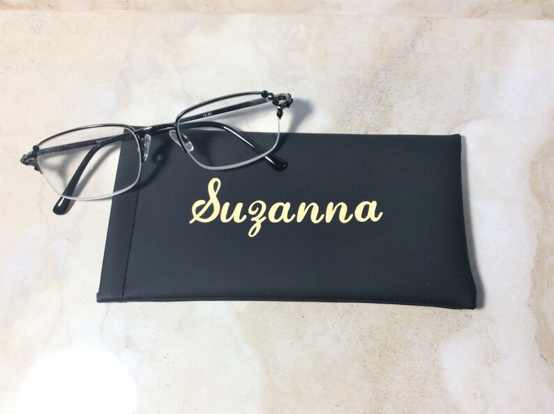 May include: Black-framed spectacles sit on a black case, personalised with the name "Suzanna" in gold script. The glasses have a half-rim design. The case is a dark rectangle.