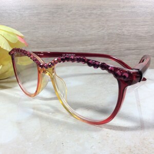 May include: Red and clear eyeglasses with a gradient lens. The top of the frames are adorned with small, purple, decorative jewels. The glasses are set against a light background with a yellow and pink flower.