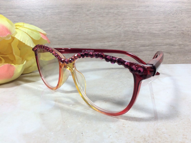 May include: Red and clear eyeglasses with a gradient lens. The top of the frames are adorned with small, purple, decorative jewels. The glasses are set against a light background with a yellow and pink flower.