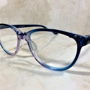 Puede incluir: Gafas con montura degradada de morado a azul. Las monturas est&aacute;n adornadas con peque&ntilde;os adornos azul oscuro. Las lentes son transparentes y las patillas son azul oscuro. El texto "UV Protection" est&aacute; impreso en la montura.