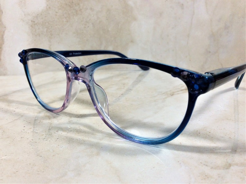 Puede incluir: Gafas con montura degradada de morado a azul. Las monturas est&aacute;n adornadas con peque&ntilde;os adornos azul oscuro. Las lentes son transparentes y las patillas son azul oscuro. El texto "UV Protection" est&aacute; impreso en la montura.
