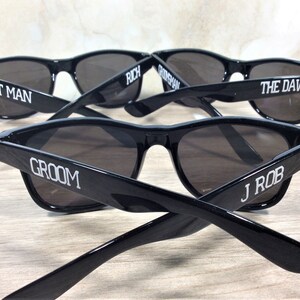 May include: Five black sunglasses with white text on the frames. The text reads "Best Man", "Groom", "Rich", "The Dawg", and "J Rob".