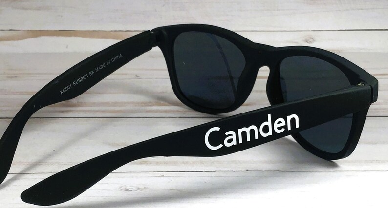 Op de afbeelding: Matzwarte zonnebril met donker getinte glazen en een klassieke montuurvorm. De buitenste poot toont de witte tekst Camden, terwijl de binnenste poot is bedrukt met de tekst KM001 RUBBER BK MADE IN CHINA in het wit.