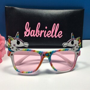 May include: White sunglasses with pink lenses and unicorn-shaped side pieces. The sunglasses have a rainbow stripe design and the text "Gabrielle" is printed on a black case.