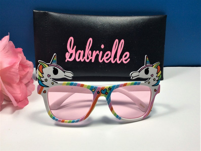 May include: White sunglasses with pink lenses and unicorn-shaped side pieces. The sunglasses have a rainbow stripe design and the text "Gabrielle" is printed on a black case.