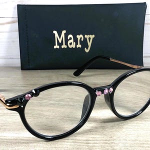 May include: Oval-framed black spectacles with pink gemstone embellishments on the frame and temples. The glasses have clear lenses and a gold-coloured arm. A black case with the name "Mary" in gold lettering is behind the spectacles.