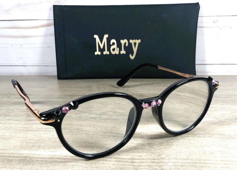 May include: Oval-framed black spectacles with pink gemstone embellishments on the frame and temples. The glasses have clear lenses and a gold-coloured arm. A black case with the name "Mary" in gold lettering is behind the spectacles.