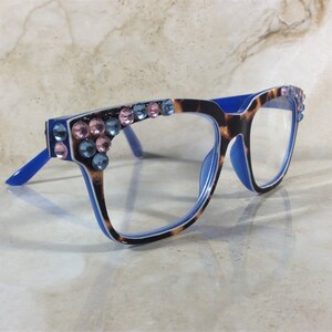 May include: Eyeglasses with a blue frame and temples, featuring a tortoiseshell pattern on the front. The frames are adorned with pink and blue rhinestones. The lenses are clear.